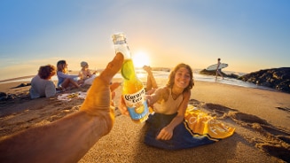 Fles Corona Cero bier met limoen op het strand, mensen ontspannen en genieten van de zonsondergang.