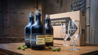 Drie flessen Tarwewijn naast twee glazen met het Hertog Jan-logo en een kartonnen doos met daarop de tekst 'Rechtstreeks uit Arcen', op een houten tafel met hopbellen.