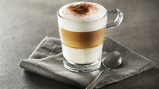 Glas latte macchiato met melkschuim en cacaopoeder, geserveerd op een grijze servet met een lepel ernaast.