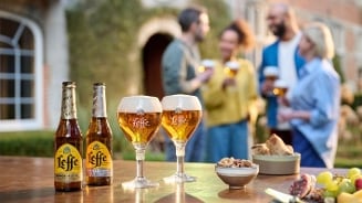 Leffe Blond bierflessen en -glazen op een tafel met snacks, met een groep mensen op de achtergrond in een tuinsetting