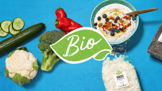 Biologisch assortiment met komkommer, rode paprika, broccoli, bloemkool, kokos snippers, chiazaad en een kom yoghurt met fruit, op een blauwe achtergrond.