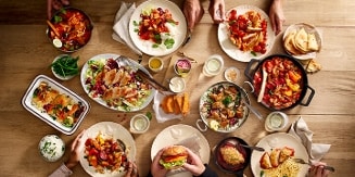 Een tafel vol diverse gerechten, waaronder salades, pasta, sandwiches, en gegrilde groenten, met meerdere handen die eten opscheppen.