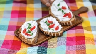 Drie sneetjes brood met roomkaas, gesneden cherrytomaten en rucola, geserveerd op een houten plank op een kleurrijk tafelkleed.