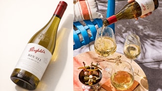 Witte wijn van het merk Penfolds wordt ingeschonken in een glas op een buitenterras, met daarnaast twee andere glazen witte wijn en een schaaltje met olijven en augurken op een ronde tafel.