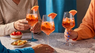 Drie mensen proosten met glazen oranje cocktail, gegarneerd met een schijfje grapefruit, aan een tafel met borrelhapjes.