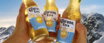 Drie flessen Corona Cero 0.0% bier worden omhooggehouden voor besneeuwde bergen onder een heldere lucht