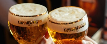 Twee glazen Grimbergen bier naast elkaar met schuimkraag, logo zichtbaar.