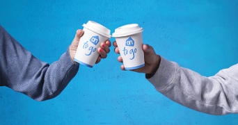 twee handen die AH to go bekers tegen elkaar tikken voor een blauwe achtergrond