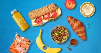 Assortiment van ontbijt en lunchproducten op een blauwe achtergrond, met onder andere een meergranenbroodje met beleg, croissant, salade met noten en groenten, banaan, appel, fles sinaasappelsap, babyworteltjes van Albert Heijn en een cappuccino