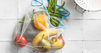 Herbruikbare zak met verschillende groenten en fruit waaronder sperziebonen, sinaasappel, avocado, banaan, peer en appel, naast een vergiet op een tegelvloer.