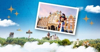 Foto van een sprookjesachtige themaparkomgeving met een groot gebouw, een persoon verkleed als figuur in kleurrijk kostuum en diverse attracties omringd door groene bomen en wolken.