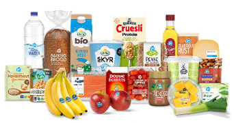 Een selectie van Albert Heijn producten, waaronder verse bananen, appels, brood, havermout, melk, skyr, pasta, rijst, water, eieren, zalm, tomaten, Douwe Egberts koffie, Quaker cruesli, pindakaas, mango, kaas, noten, olie en prei, uitgestald op een witte ondergrond.