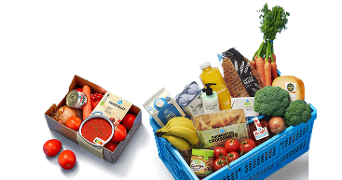 Blauwe Albert Heijn winkelmand gevuld met diverse boodschappen zoals vers brood, melk, bananen, broccoli, wortels, sinaasappelsap, tomaten, croissants, kaas, yoghurt en handzeep, naast een doosje met ingrediënten voor tomatensoep zoals tomaten, ui en wortel.