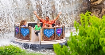 Groep mensen in een ronde boot beleeft plezier tijdens een wildwaterattractie in een pretpark, met opspattend water en een waterval op de achtergrond.