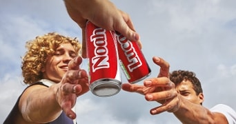 Twee personen reiken naar rode Noah energiedrank blikjes onder een blauwe lucht.