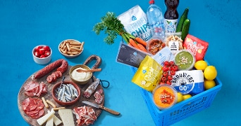 Mandjes gevuld met AH producten, zoals wortelen, tomaten, citroenen, AH Robuuste Chips, AH Oven Toppers, en Brouwers 0.0% bier, naast een houten plank met diverse hapjes zoals kaas, ansjovis, en worst.