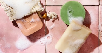 Verschillende stukken zeep, waaronder een roze, een groene en een witte, op roze tegels met schuim.