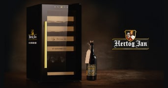 Zwarte koelkast met het Hertog Jan logo naast een fles Hertog Jan bier en een label, op een donkere achtergrond met het Hertog Jan logo prominent in beeld.