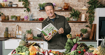Persoon in keuken met open kookboek van Jamie Oliver tussen verse groenten en fruit