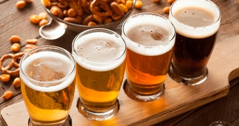 Vier glazen bier in verschillende kleuren op een houten plank, met gezouten noten en zoutjes op de achtergrond.