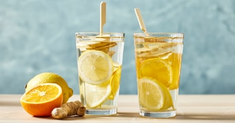 Twee glazen met water, citroenschijfjes, gember en sinaasappel op een houten tafel. Op de voorgrond liggen een halve sinaasappel, een citroen en verse gember.
