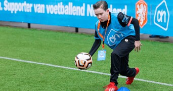 Kind speelt voetbal op grasveld met AH-logo hesje en oranje schoenen.