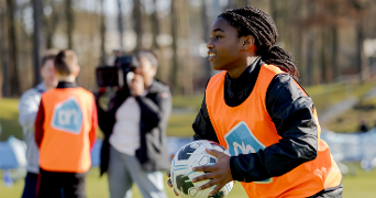 Meisje in een voetbaltraining met een oranje AH-hesje en een voetbal, op een sportveld met andere deelnemers en een cameraman op de achtergrond.