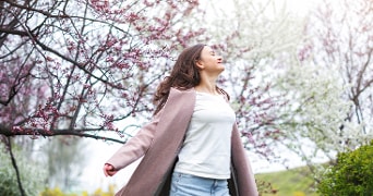 Vrouw in een lentetuin met bloeiende bomen, gekleed in een lichtroze jas en witte t-shirt, genietend van de buitenlucht.