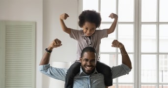 Kind met opgetrokken armen zit op de schouders van een volwassene in een lichte kamer.