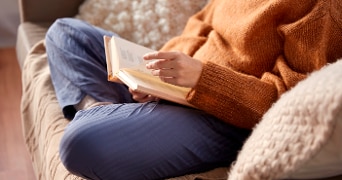 Persoon in pyjamabroek en oranje trui een boek aan het lezen op de bank