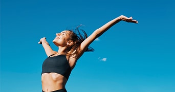 Vrouw in sportkleding steekt haar armen in de lucht tegen een blauwe hemel