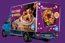 Albert Heijn bestelwagen met kerstversiering en Allerhande tijdschrift, inclusief een bord met feestelijke hapjes en de tekst 'Kerst loopt op rolletjes' op een paarse achtergrond