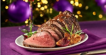 Rosbief in plakken gesneden op een wit bord, geserveerd met geroosterde rode ui en knoflook, op een paarse ondergrond met feestelijke verlichting op de achtergrond