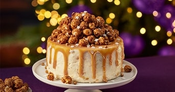 Karameltaart met witte glazuur, gegarneerd met gezouten karamelpopcorn en parelsuiker, op een feestelijk gedekte tafel met kerstverlichting op de achtergrond