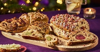 Verschillende kerststollen met vullingen zoals amandelspijs en noten, gesneden en gepresenteerd op een houten plank, met feestelijke achtergrondverlichting.