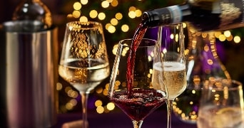 Rode wijn wordt ingeschonken in een wijnglas naast glazen met witte wijn en mousserende wijn, op een feestelijke achtergrond met lichtjes