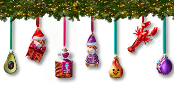 Kerstversiering in de vorm van grappige ornamenten, waaronder een avocado, beertje met cadeautje en kerstmuts, AH volkoren brood, beertje met trui, lachende peer, kreeft en een macaron, hangend aan linten onder een kersttak met lichtjes