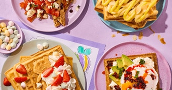 Vier borden met verschillende soorten wafels, elk creatief belegd met toppings zoals aardbeien en slagroom, tomaat en feta, avocado en ei, en banaan met amandelen, op een paarse ondergrond met een schaaltje gekleurde snoepjes ernaast.