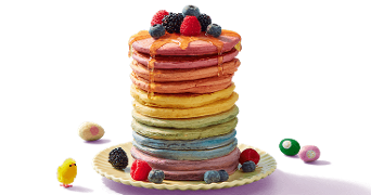 Stapel regenboogpannenkoeken met honing en vers fruit, zoals aardbeien, bramen en bosbessen, op een bord, omringd door kleine paaseitjes en een decoratief geel kuikentje.