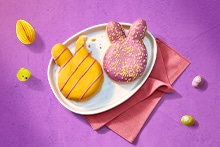 Verschillende kleurrijke paastraktaties op een tafel, waaronder koekjes, chocolade-eitjes, paashaashapjes, macarons en een chocolade paashaas, geserveerd op borden en schaaltjes.