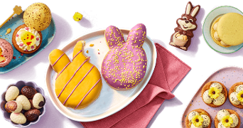 Verschillende kleurrijke paastraktaties op een tafel, waaronder koekjes, chocolade-eitjes, paashaashapjes, macarons en een chocolade paashaas, geserveerd op borden en schaaltjes.