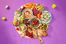Borrelplank met diverse hapjes waaronder hummus, tapenade, druiven, olijven, groente, kaasblokjes, crackers, wraps en vleeswaren.