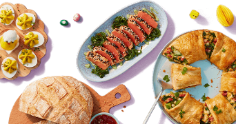 Diverse paasgerechten op tafel, waaronder gevulde eieren, een schaal met sesamtonijn op groenten, krokant brood en een hartige taartkrans.