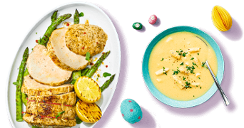 Gesneden varkenshaas met gegrilde asperges en citroen op een schaal, naast een kom romige aspergesoep en paasdecoratie.