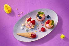 Assortiment van kleurrijke paasdesserts, waaronder wafels met slagroom en aardbeien, meringues met fruit, mini taartjes met bessen, en een hoorntje met framboos, gepresenteerd op verschillende borden. Paasmaker logo linksonder.