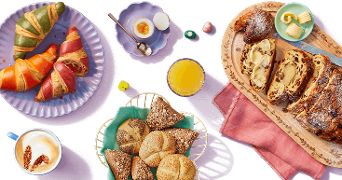 Ontbijttafel met verschillende soorten broodjes, croissants, gesneden brood met rozijnen, een gekookt ei, sinaasappelsap, cappuccino en boter.