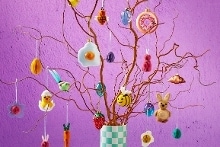 Paastak met decoraties zoals konijnen, paaseieren, bloemen, een friet ei en kleurrijke ornamenten in een geruite vaas tegen een paarse achtergrond