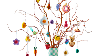 Paastak met decoraties zoals konijnen, paaseieren, bloemen, een friet ei en kleurrijke ornamenten in een geruite vaas tegen een paarse achtergrond