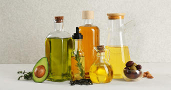 Verschillende glazen flessen gevuld met plantaardige oliën, omringd door een halve avocado, olijven, noten en kruiden op een lichte achtergrond.