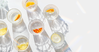 Zeven glazen water met schijfjes citrusfruit, waaronder sinaasappel en citroen, op een witte ondergrond met opvallende lichtreflecties.